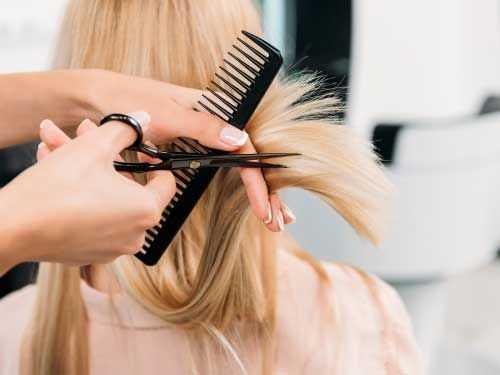 Hairdresser cutting a client's blonde hair using scissors and a comb in a salon.